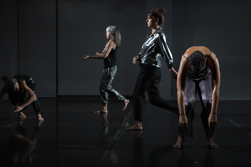 Two women in metallic walk in profile. In the foreground, one woman is flopped over, her arms dangling to the ground. To the left, is a woman crouched on the floor.